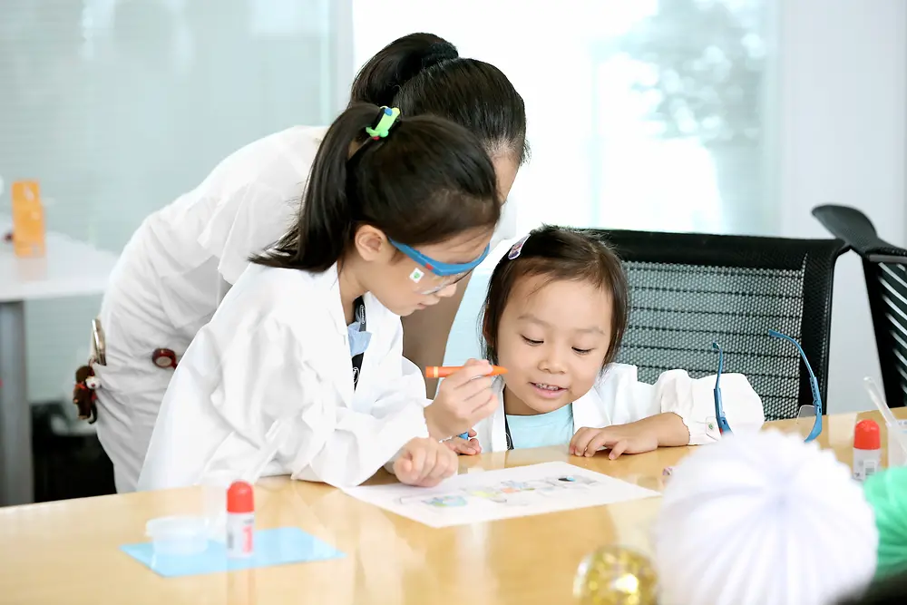 Двоє дітей і жінка в лабораторному халаті розфарбовують малюнок за столом