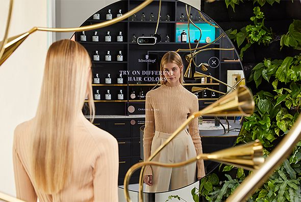 A blonde woman is standing in front of a round mirror and looks at her reflection. The interior of the House of Schwarzkopf is mirrored, too.