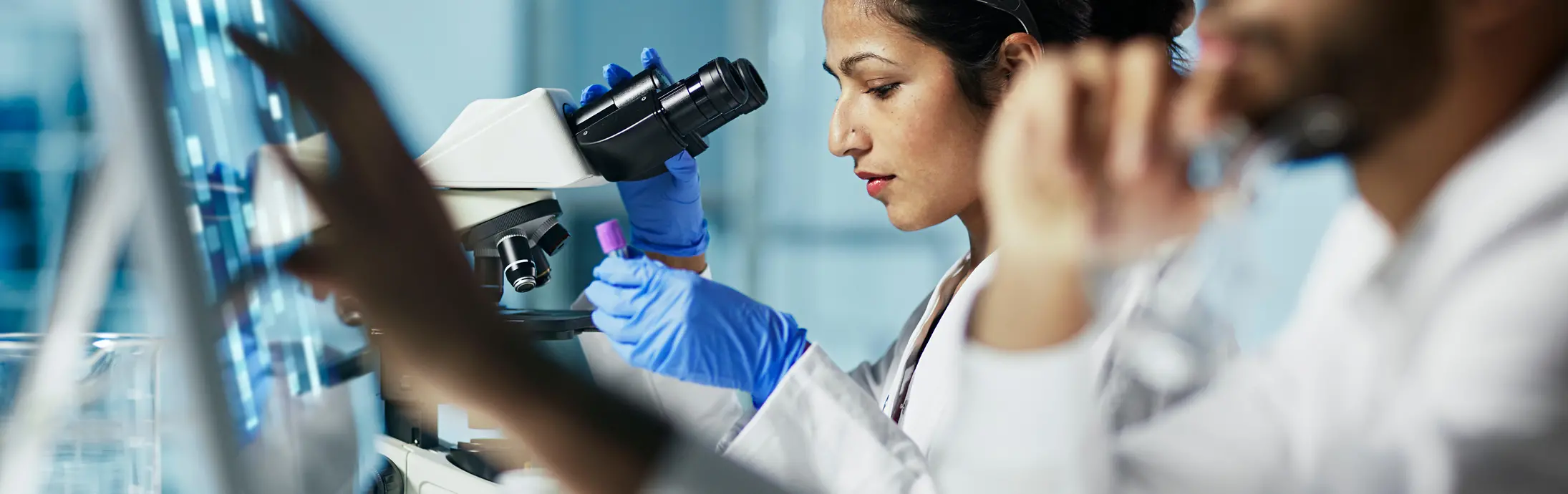 жінка сидить у лабораторії перед мікроскопом, розглядаючи зразок, поруч неї чоловік з бородою, котрий дивиться на екран 