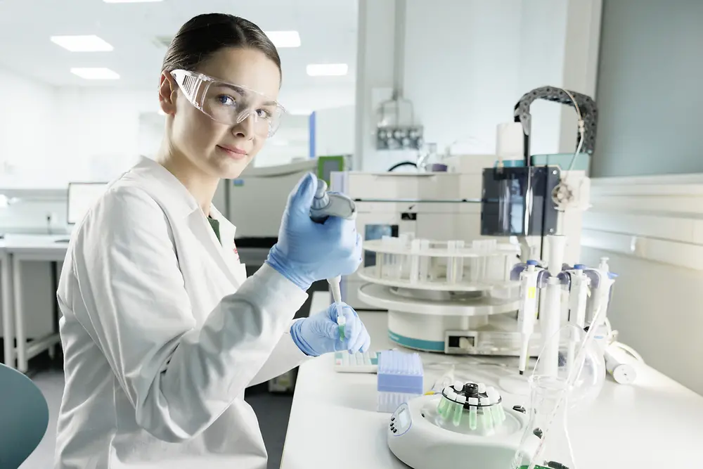 Жінка-науковиця експериментує в лабораторії, одягнена в лабораторний халат, на обличчі – захисні окуляри.