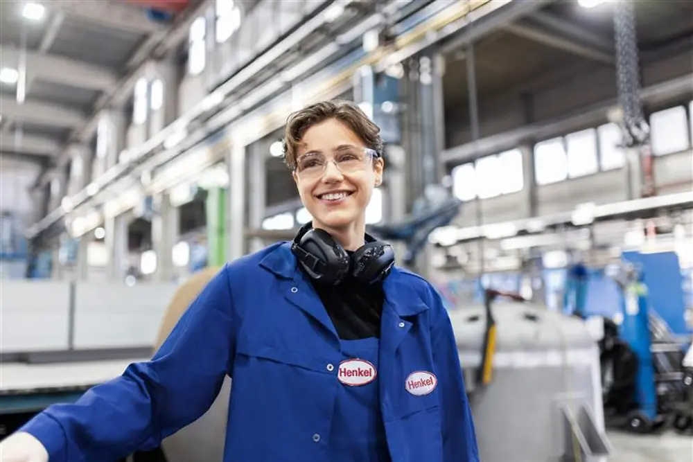Усміхнений співробітник Henkel у синьому робочому халаті та захисних навушниках стоїть у виробничому цеху, уособлюючи професіоналізм та інновації у сфері.