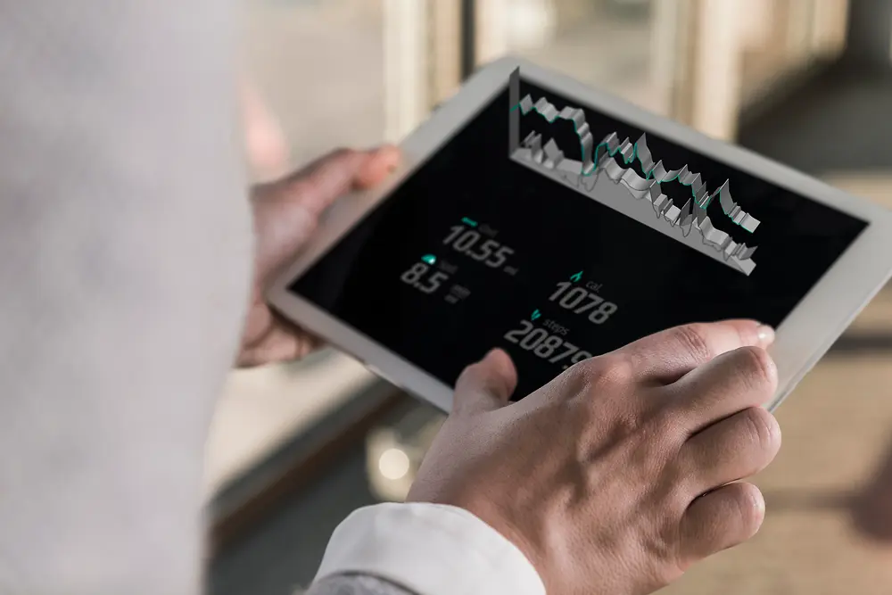 Людина тримає планшет, на якому відображаються цифрові діаграми та числові дані; вигляд через плече у світлому приміщенні.